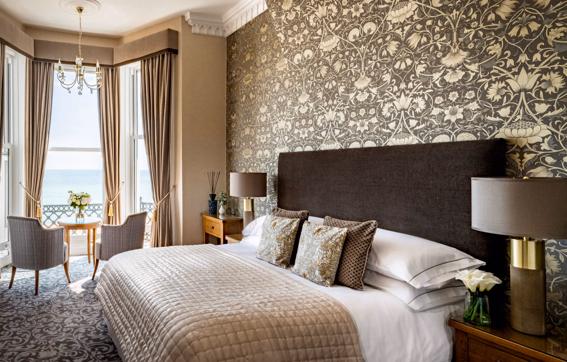 A large bed in a room with a sea view, a table and chairs and bedside table